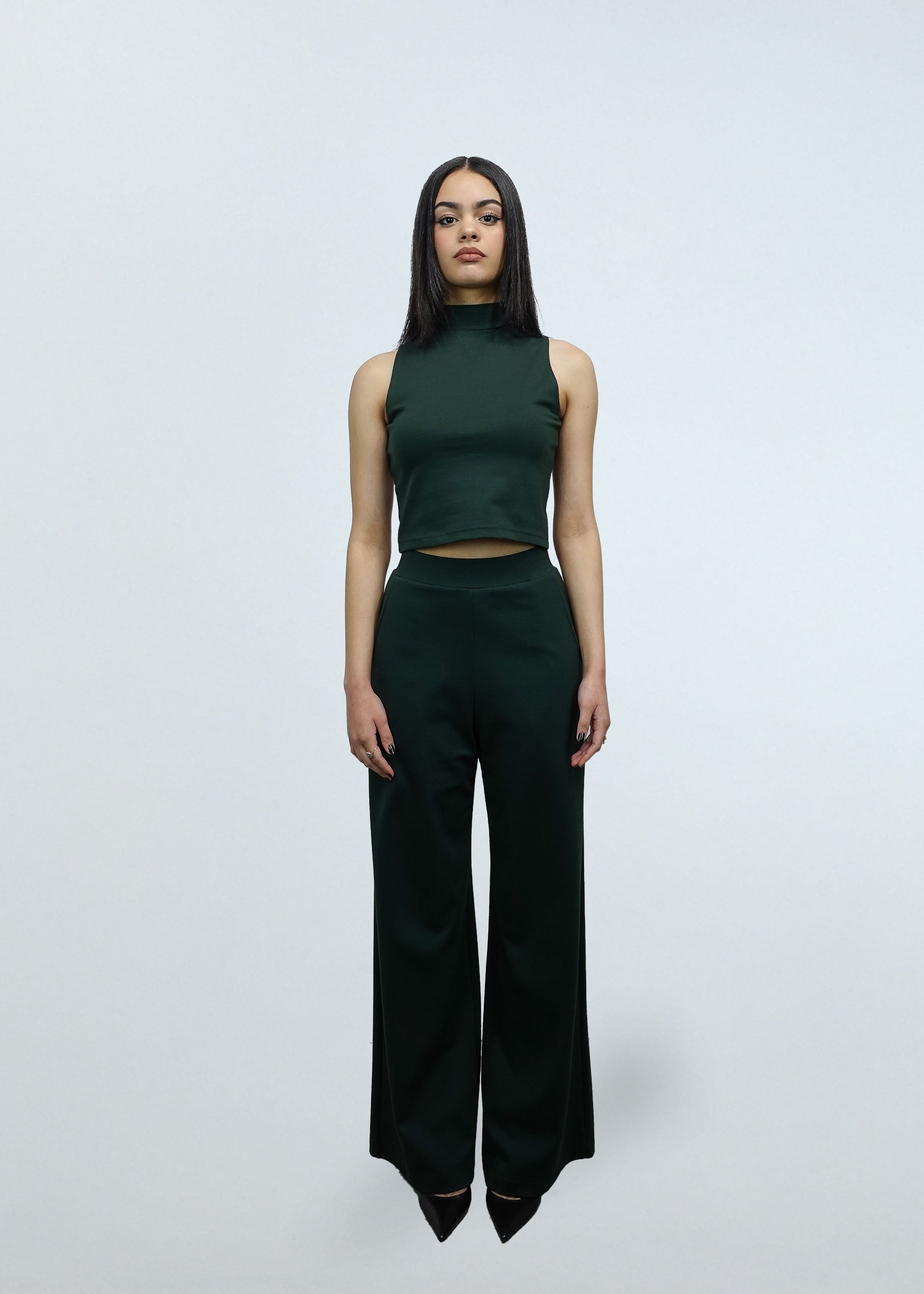 Woman wearing a green sleeveless top and dark green high-waisted pants on a white background